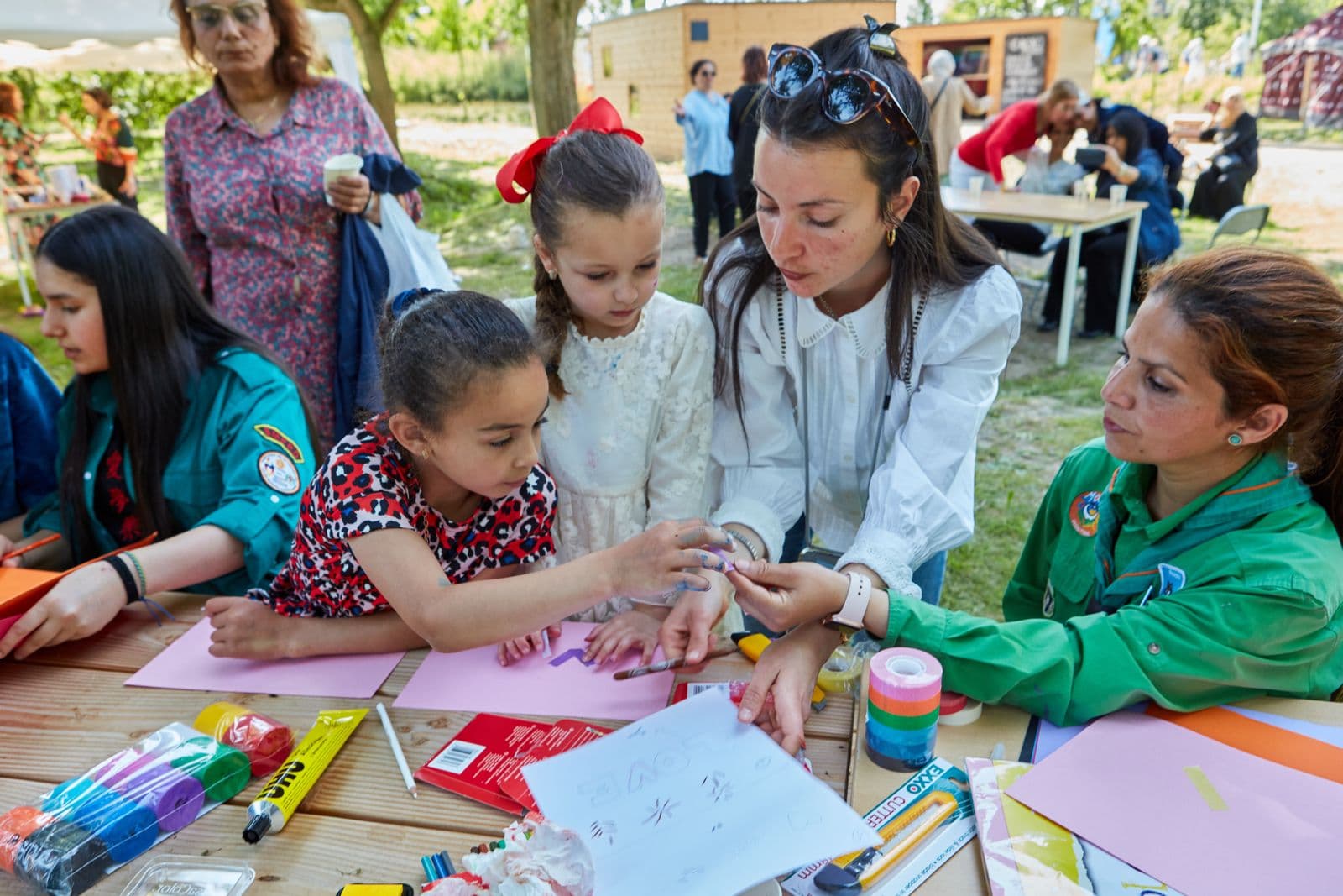 Buitenactiviteit: kinderen, vrijwilligers en leiding in scoutinguniform rond een tafel met knutselen en tekenen op gras, zonnige dag